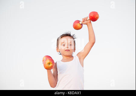 handsome caucasian little boy lifting overhead dumbbells made from red apples on bright background Stock Photo