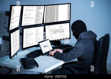 Male hacker using laptop against multiple monitors at desk in office Stock Photo