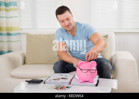 Mid adult man putting coin into piggy bank while calculating finance at home Stock Photo