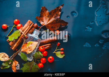 Glass perfume bottle with fallen maple leaves, red berries, cinnamon and autumn rain on a wet wooden background Stock Photo