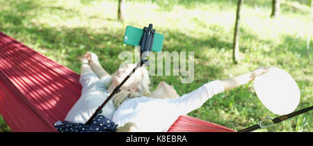Young friends in sweaters making selfie photo with smart phone sitting together on the hammock in the garden Stock Photo