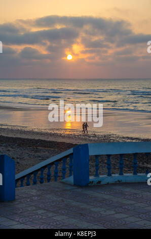 Two locals walking along beach promenade with stone walkway during sunset at Sidi Ifni, Morocco, North Africa. Stock Photo