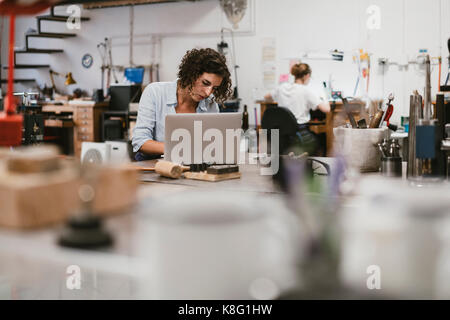 Female jeweller typing on laptop at workbench Stock Photo
