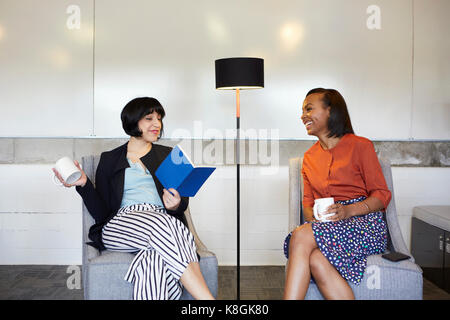 Two businesswomen sitting in comfortable chairs in office, having coffee break Stock Photo
