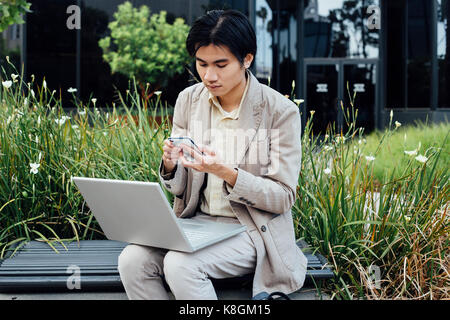 Young man sitting outdoors, using laptop, holding smartphone Stock Photo