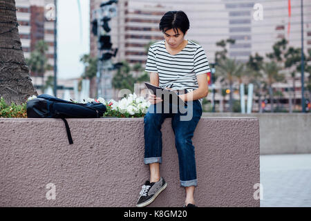 Young man sitting on wall, outdoors, writing in notebook Stock Photo