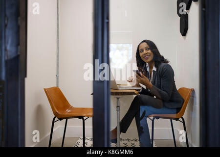 Businesswoman sitting in office, using laptop, holding smartphone, smiling Stock Photo