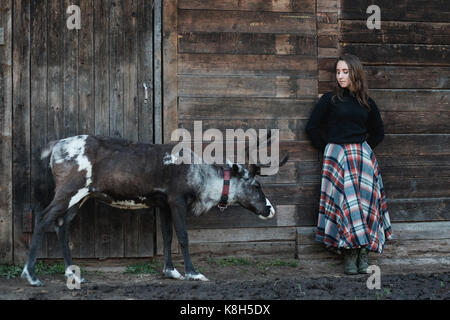 A young European girl in a plaid skirt is standing next to a reindeer near a wooden wall. Stock Photo