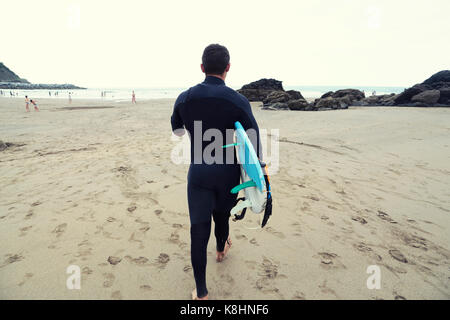 Rear view of man in wetsuit carrying surfboard while walking on beach Stock Photo