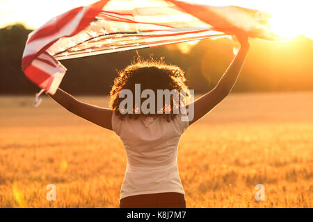 Rear view of mixed race African American girl teenager female young woman in a field holding USA stars and stripes flag above her head in golden sunse Stock Photo