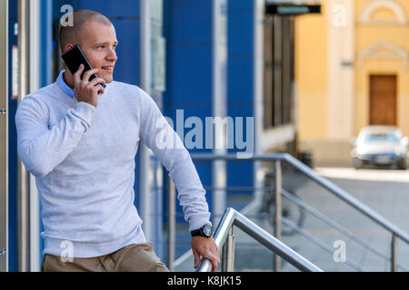 Young handsome businessman standing in front of the building and talking on a mobile phone. He arranges a meeting with his new business partners via a Stock Photo