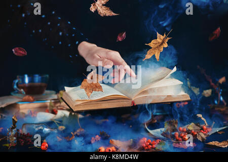 Still life with levitating book of spells, autumn leaves, red berries, jars and bottles on a dark background with rising mystic smoke and a hand in a black shirt with star print Stock Photo