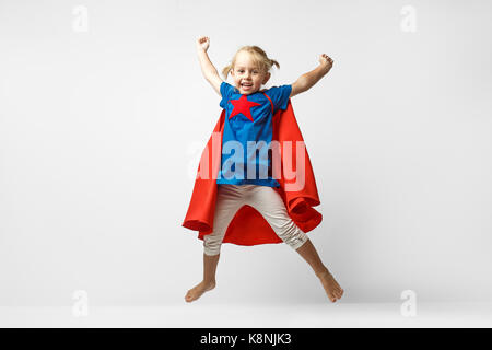 Very excited little girl dressed like hero jumping alongside the white wall. Stock Photo