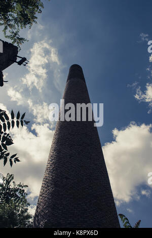 Tall old brick industrial chimney rising against a blue sky with white clouds, framed by tree foliage, low angle perspective view Stock Photo