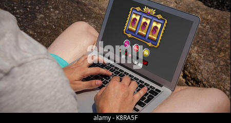 Slot Machine on mobile display against man sitting on the rocks and using laptop at the sea coast Stock Photo