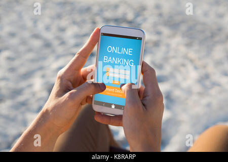 Online banking text on blue mobile screen against hands of man using mobile phone at beach Stock Photo