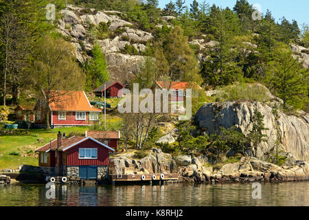 Rock coast along a Norwegian fjord with red painted wooden vacation houses on a hillside. Stock Photo