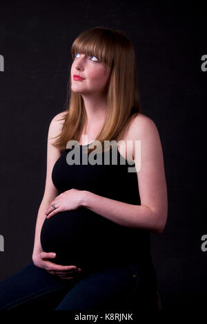 Portrait of a pregnant woman holding her belly with a thoughtful expression on her face. Portrait shot with black background. Stock Photo