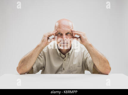 Contemplative balding mature man sitting at table against plain background Stock Photo