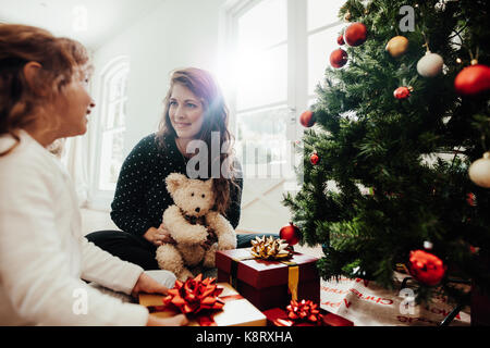 Mother with her child daughter celebrating Christmas. Young woman with her daughter sitting beside Christmas tree holding gift boxes. Stock Photo