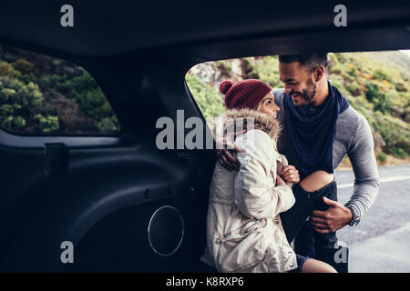 Young couple in sitting together in the car trunk on the roadside. Romantic man and woman traveling by car. Stock Photo