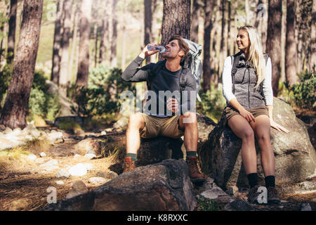Man and woman hikers relaxing while trekking in forest. Hiker couple taking a break sitting on rock and drinking water. Stock Photo