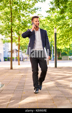A handsome young businessman walking on the sunny street surrounded by trees while holding his laptop and talking on a phone Stock Photo