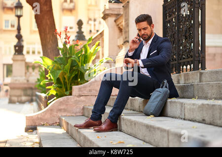 A handsome young businessman sitting on the stairs talking on his smartphone and checking the time on his watch at the Basilica in Budapest Hungary. Stock Photo