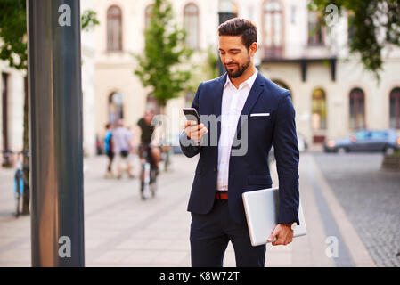 A handsome young businessman holding his laptop while using his phone and standing on the street Stock Photo