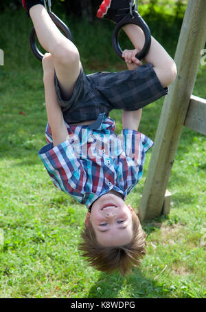 a smiling brown haired boy grins as he hangs upside down on rings on a swing set outside Stock Photo