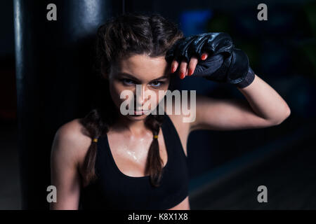 Young fighter boxer fit girl wearing boxing gloves tired after training with heavy punching bag in gym. Low key image. Woman power Stock Photo