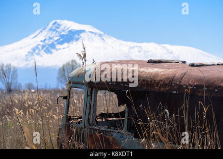 Abandoned and rusty old Soviet Russian bus in the middle of reeds and agriculture fields with snow-capped scenic Ararat mountain and clear blue sky on Stock Photo