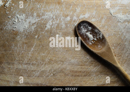 Overhead shot of a used and scratched old wooden chopping board scattered with white flour. A wooden spoon rests diagonally on its surface. Stock Photo