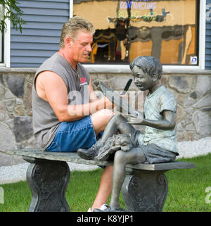 Man sitting on bench next to statue of boy reading a book texting on a cell phone Stock Photo