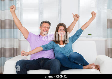 Excited Couple Raising Hands While Sitting On Sofa At Home Stock Photo