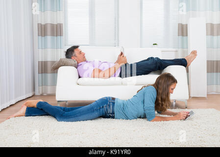 Mature Man And Woman With Digital Tablets Lying On Sofa And Carpet In Living Room Stock Photo