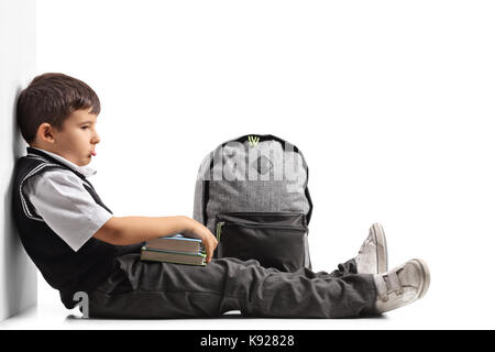 Sad schoolboy with a backpack and books seated on the floor isolated on white background Stock Photo