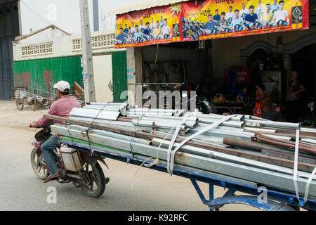 A man driving a motorcycle with an attached trailer loaded with metal pipes. Driving through the busy roads of Phnom Penh, Cambodia, South East Asia Stock Photo