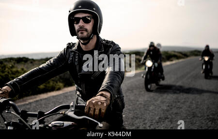 Man wearing open face crash helmet and sunglasses riding cafe racer motorcycle along rural road. Stock Photo