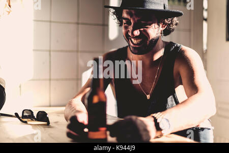 Bearded man wearing hat sitting indoors at a table, beer bottle in foreground. Stock Photo