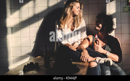 Man with brown hair holding beer bottle and a woman with long blond hair sitting indoors, looking at each other, smiling. Stock Photo