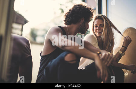 Man with brown hair wearing ripped jeans and a woman with long blond hair sitting outdoors, looking at each other, smiling. Stock Photo