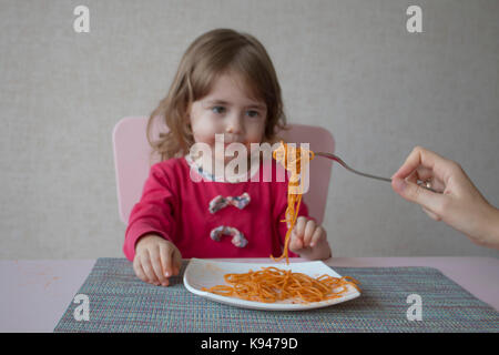 mom feeds her daughter's spaghetti. Little girl eat pasta in the kitchen table Stock Photo