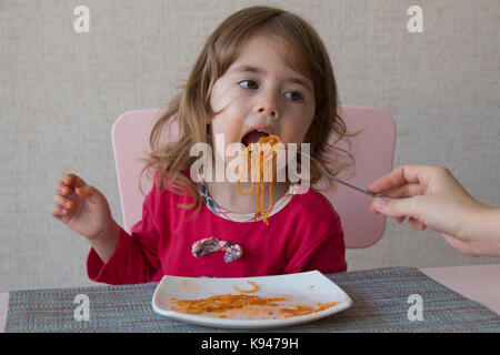 mom feeds her daughter's spaghetti. Little girl eat pasta in the kitchen table Stock Photo