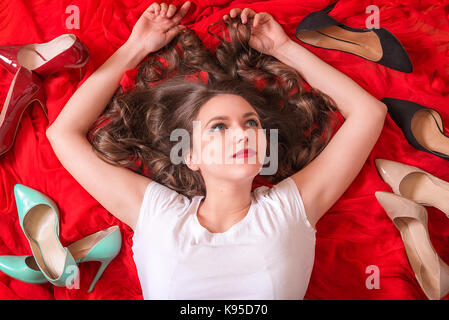 Beautiful woman with blue eyes and red lips, lays on a red fabric, smiling and surrounded by many elegant shoes in a variety of colors. Stock Photo