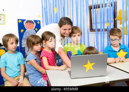 Educator surrounded by  kids at kindergarten using a laptop Stock Photo