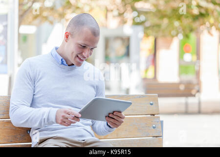 Handsome young smiling  man, student, sitting on a bench in the city and uses digital tablet Stock Photo
