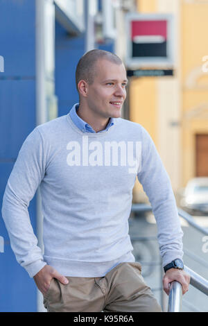Handsome and attractive young man in a shirt  leaning against the metal fence and waiting for someone. Portrait of an urban  and smiling young man out Stock Photo
