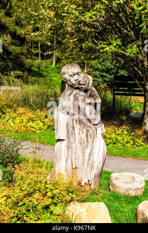 A wooden tree trunk carved into a sculpture of a woman holding a baby, lit by autumn sunlight in the forested area of Queen's View visitor centre Stock Photo