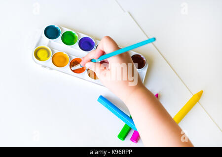 The child holds the brush over the palette and ready to paint on a blank sheet of white paper with watercolors Stock Photo